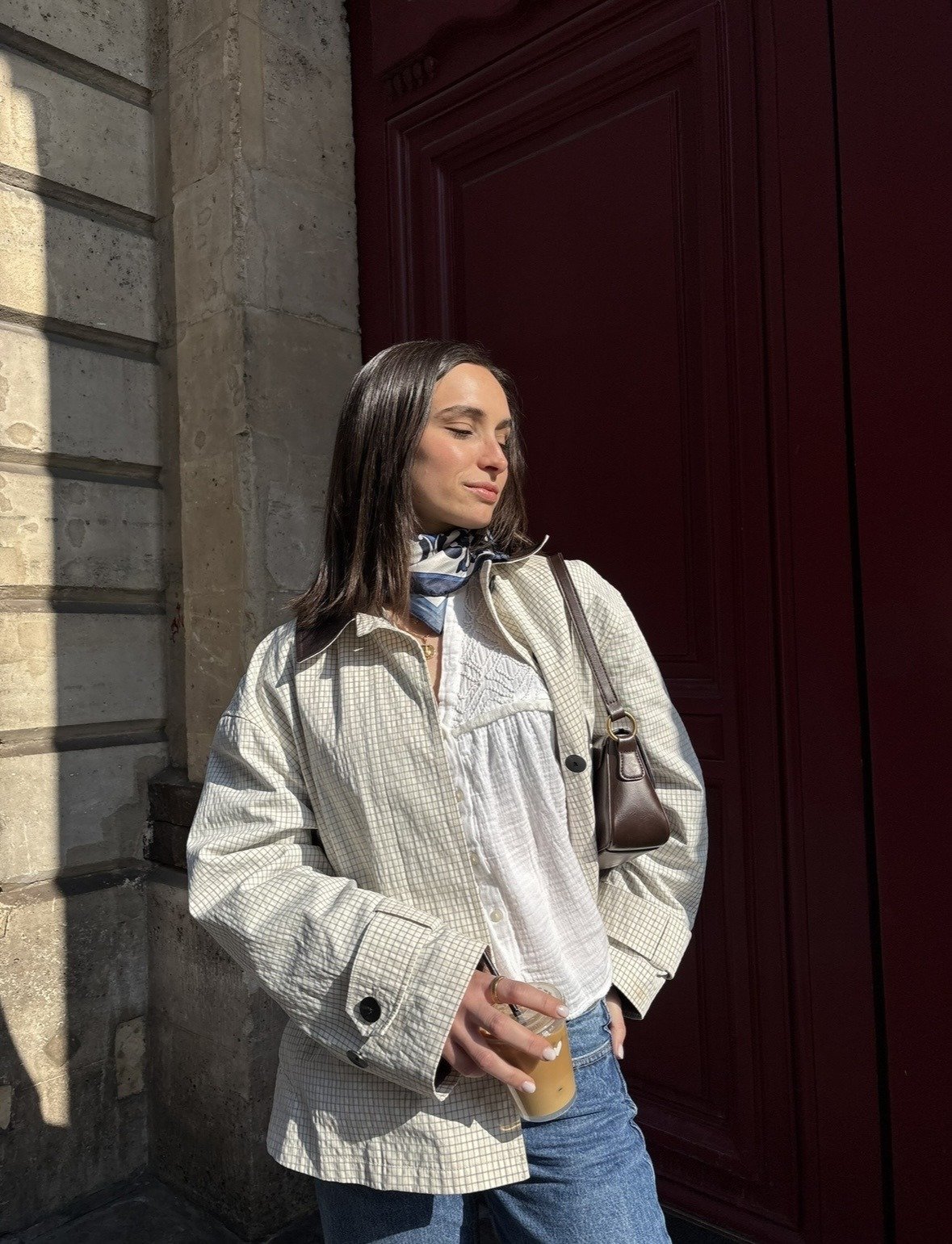 Oversized light checked jacket layered over a white textured blouse, blue jeans, a neck scarf, and a dark brown shoulder bag.