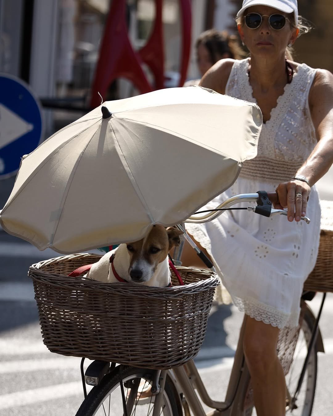 A white sleeveless eyelet dress is styled with a white cap, round sunglasses, and metallic wristwatch.