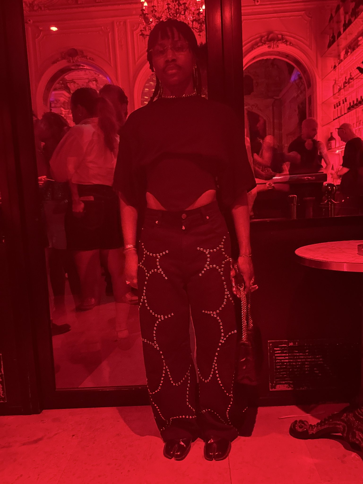 A dark fitted top with waist cutouts is styled with studded black wide-leg pants, glossy split-toe shoes, glasses, and metallic jewelry.