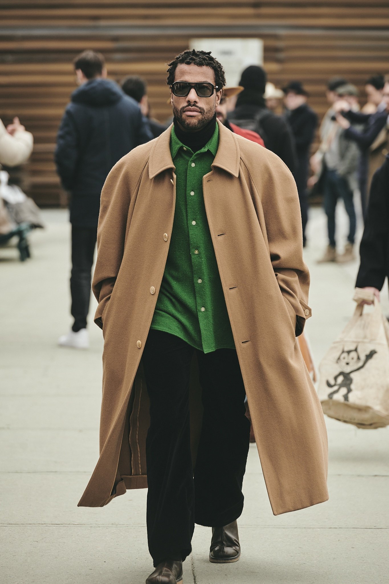 A camel overcoat is worn open over a green button-up shirt, black high-neck layer, black wide trousers, dark leather boots, and oversized rectangular sunglasses.
