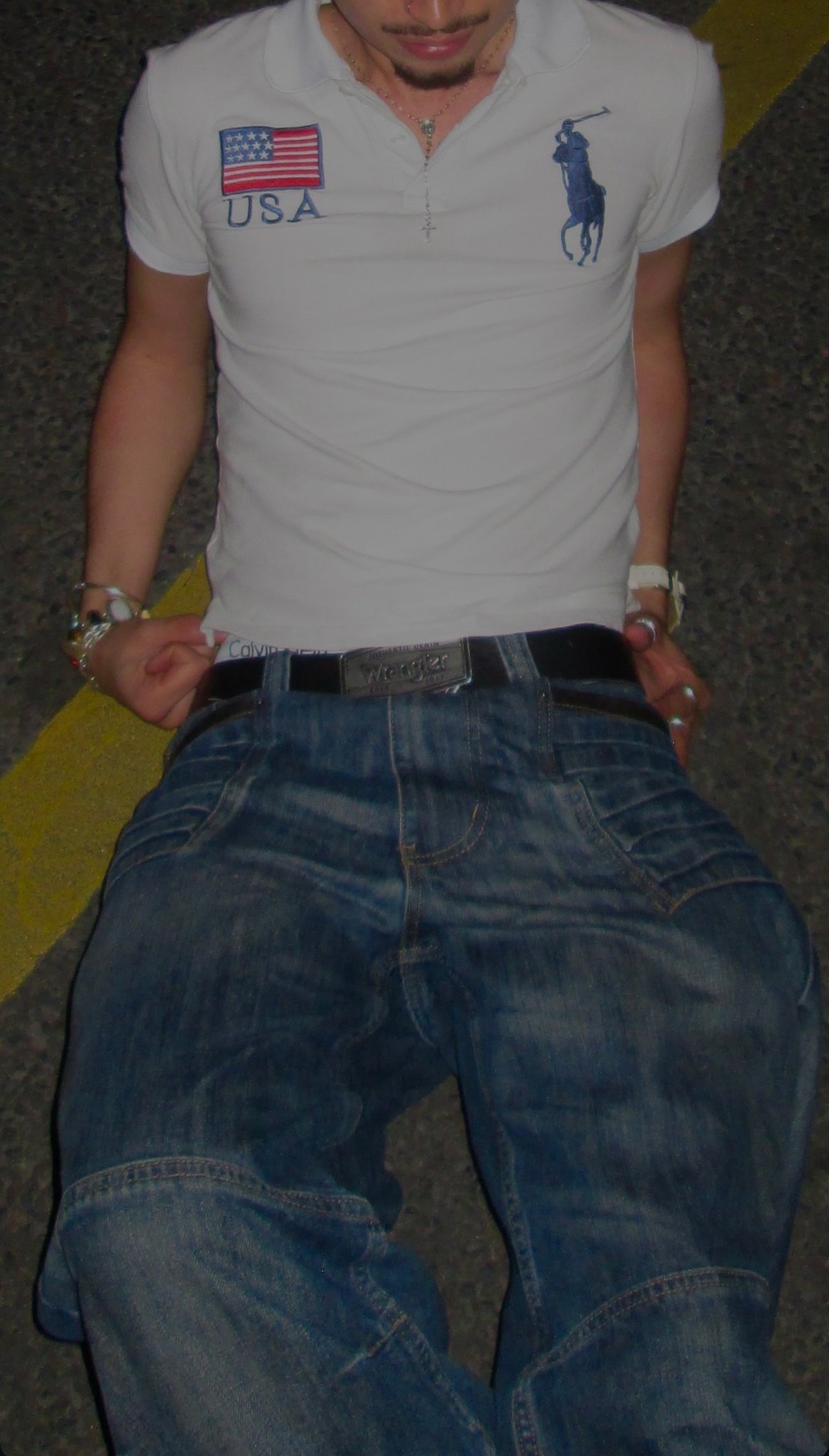 A white logo polo is worn with faded blue jeans, a black belt, layered necklaces, rings, bracelets, and a white wristwatch.