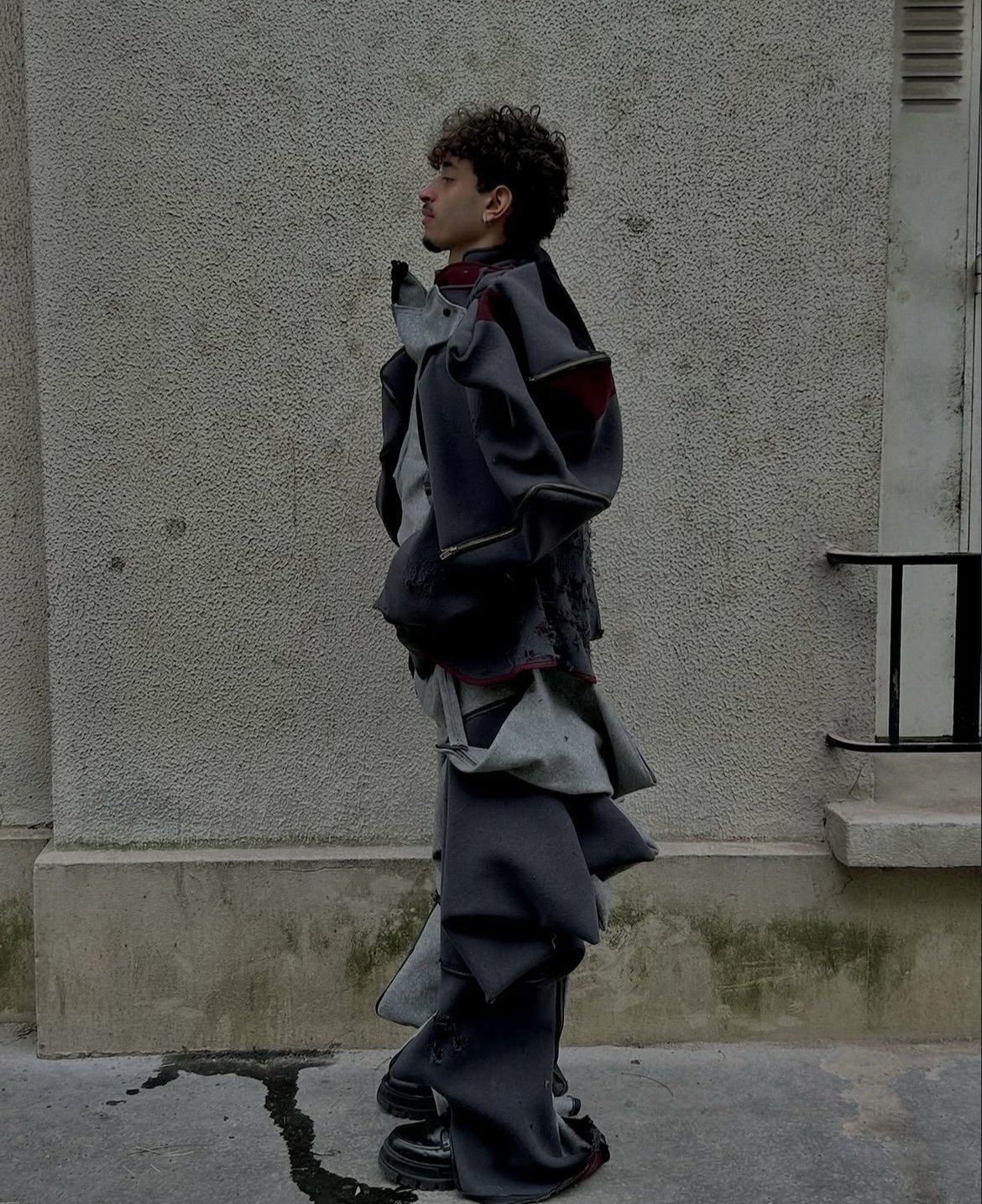 A layered grey and charcoal sculptural look with oversized folded panels and black chunky boots.