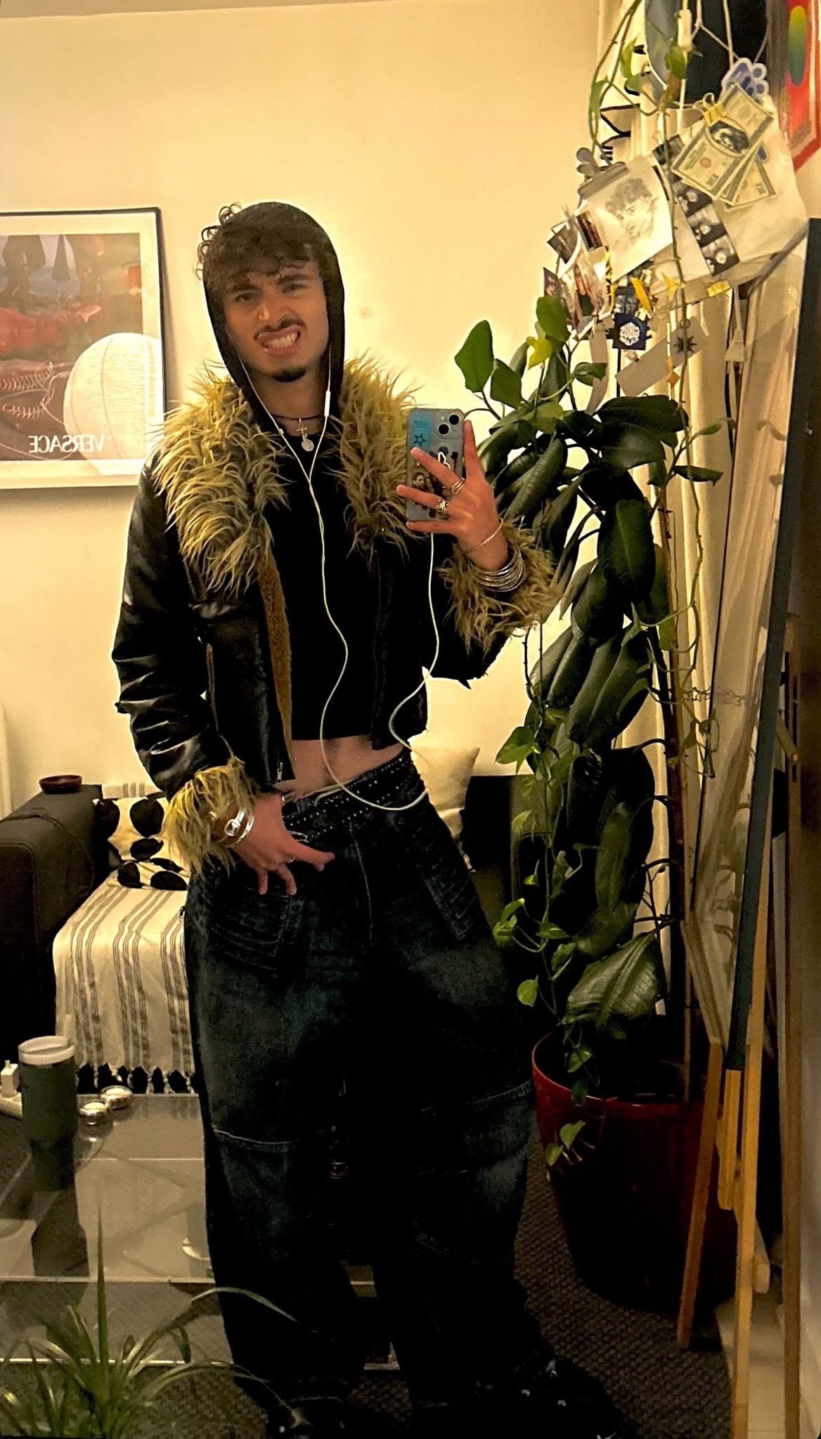 A black cropped hooded top sits under a black jacket with shaggy tan faux-fur trim, styled with wide dark denim pants, layered jewelry, and wired earphones.