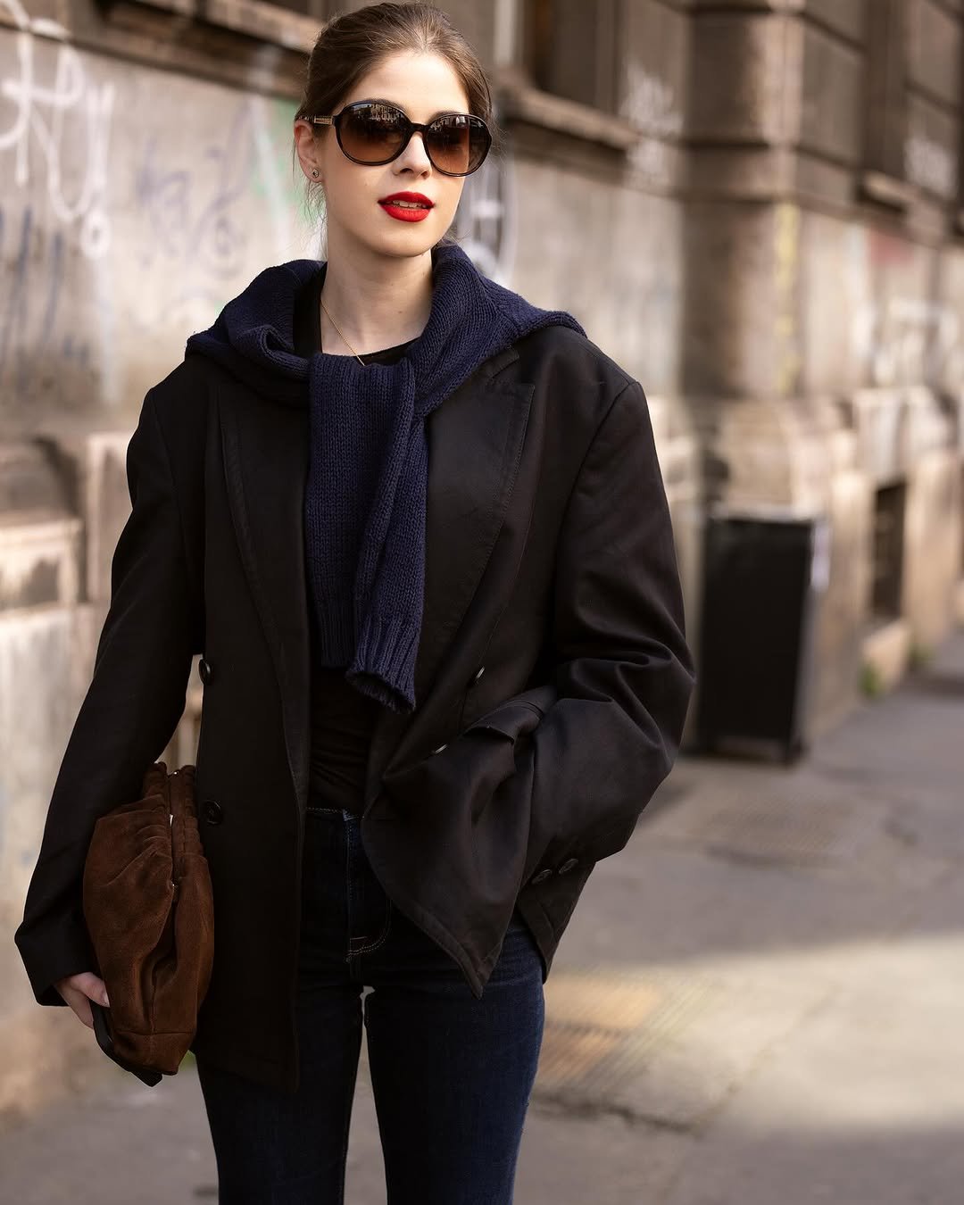 A black oversized blazer is styled over dark denim with a navy knit sweater tied around the shoulders and a brown suede shoulder bag.
