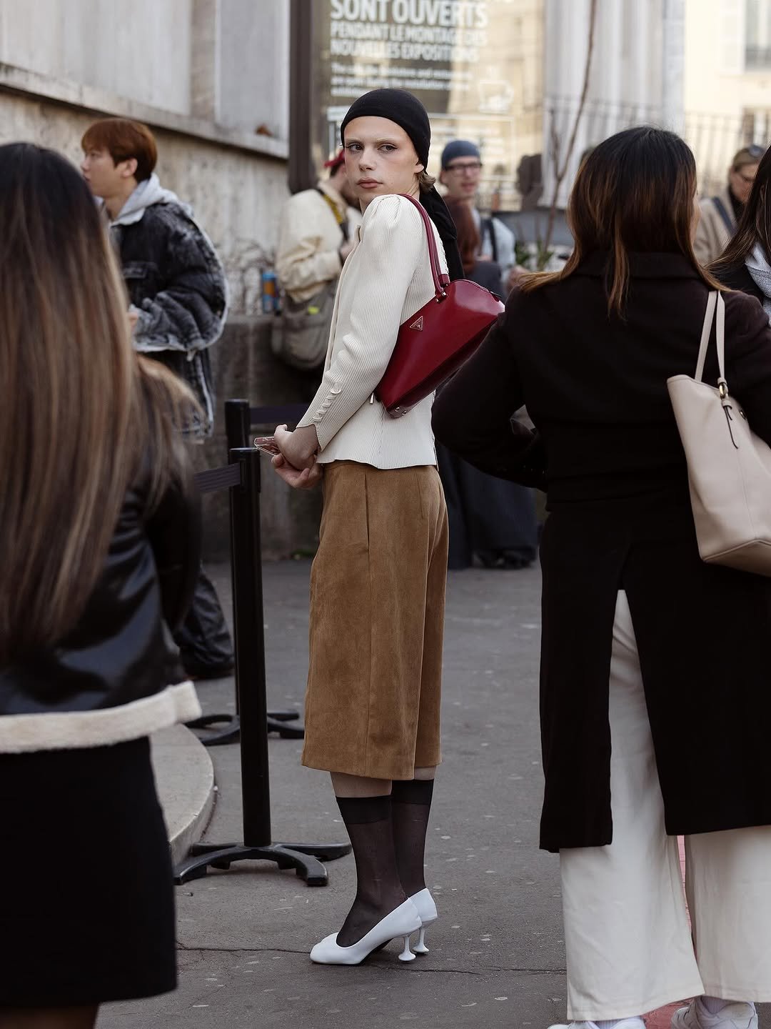 A cream ribbed cardigan, tan knee-length skirt, sheer black socks, white pumps, and a red shoulder bag create a polished street-style look.