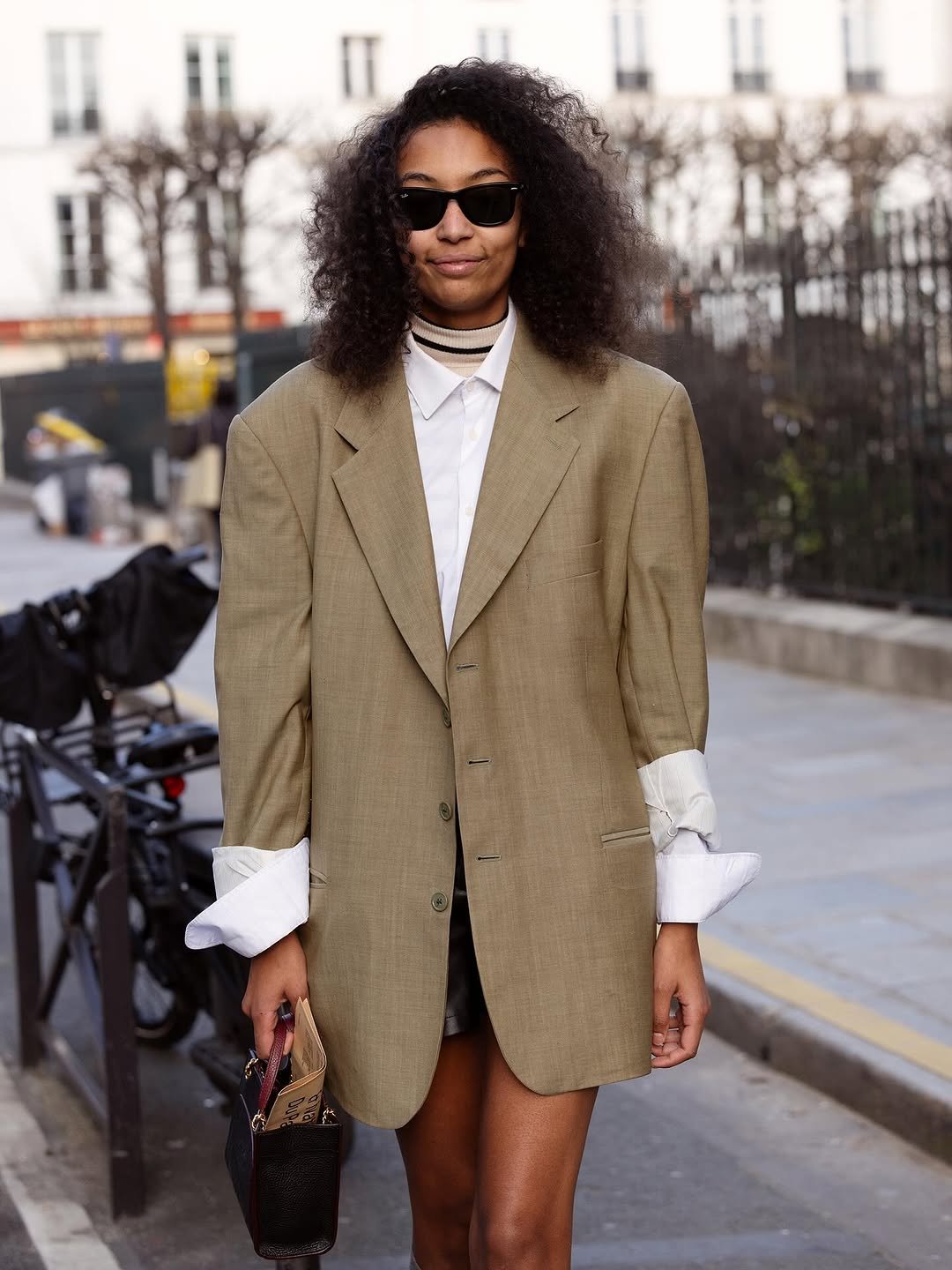 An oversized tan blazer is layered over a white shirt with dramatic folded cuffs, black mini bottoms, and a small black top-handle bag.