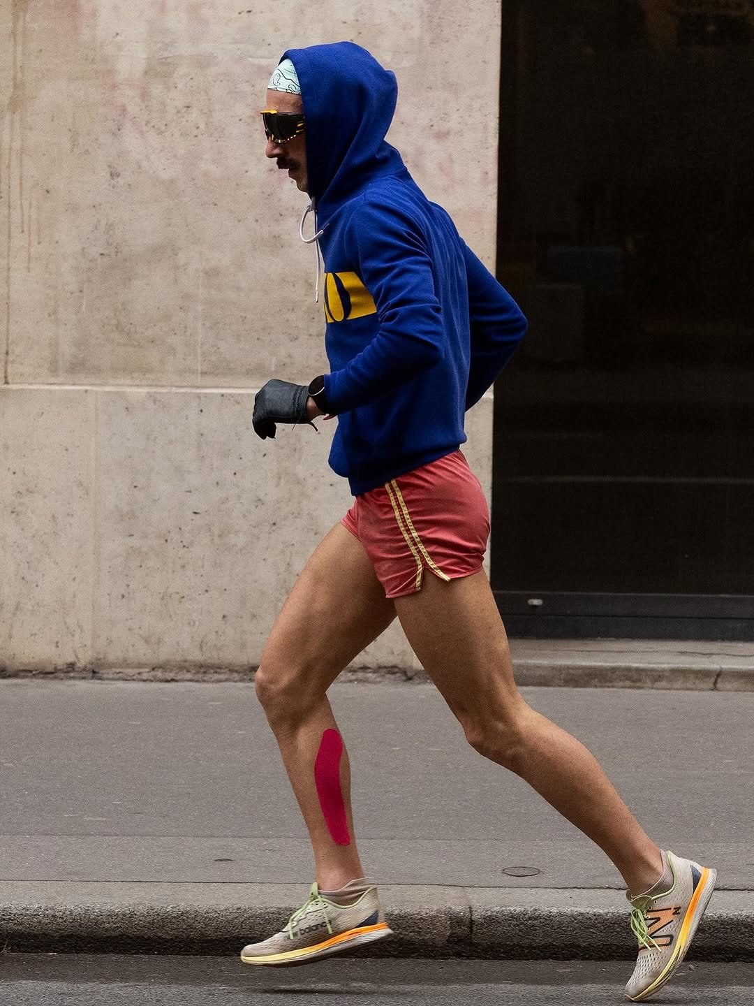 A runner wears a royal blue hooded sweatshirt, red split running shorts, performance sneakers, sunglasses, gloves, and a patterned bandana.