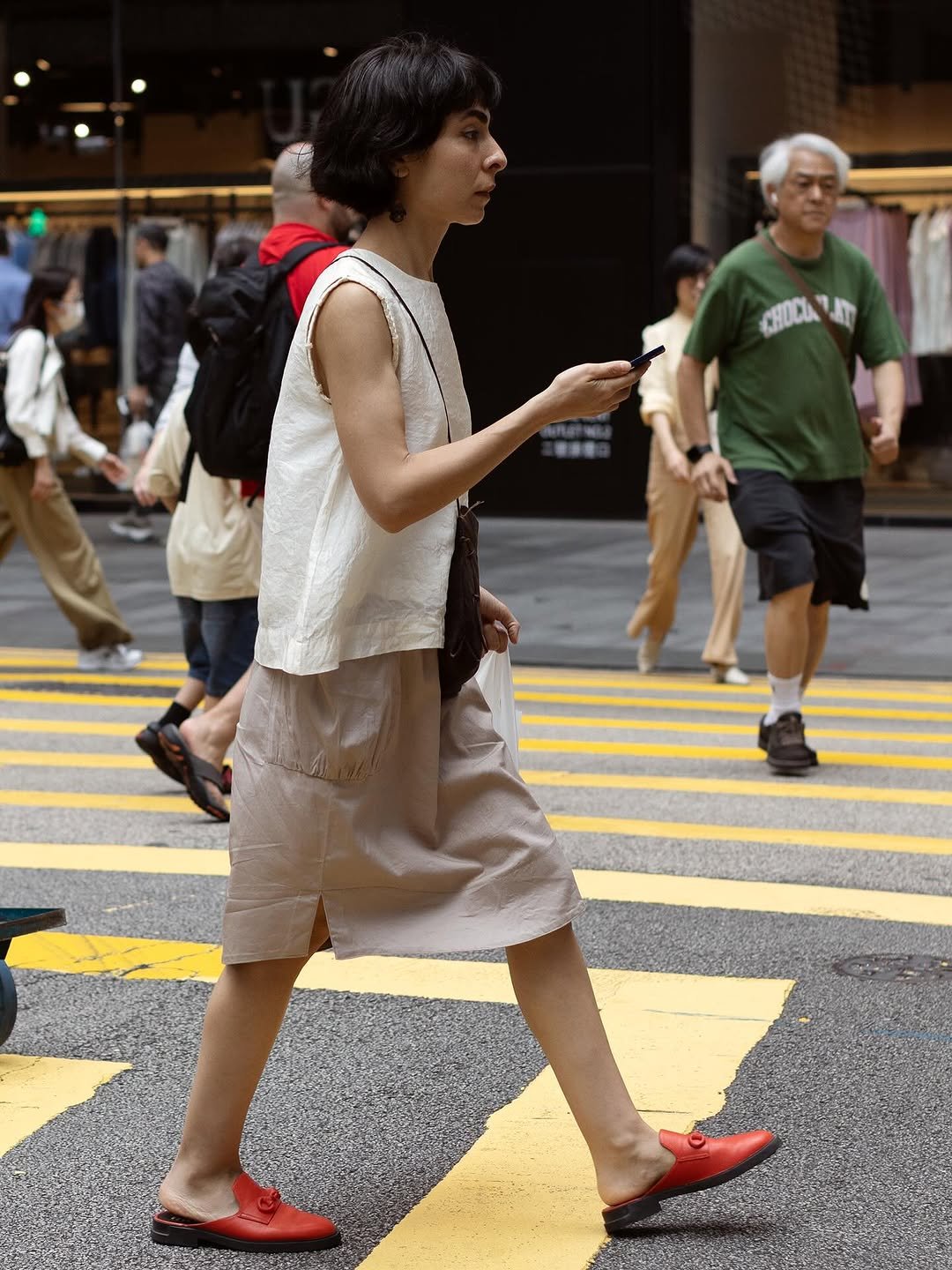 Sleeveless cream top over a taupe knee-length skirt, finished with a small dark crossbody bag and red backless loafers.