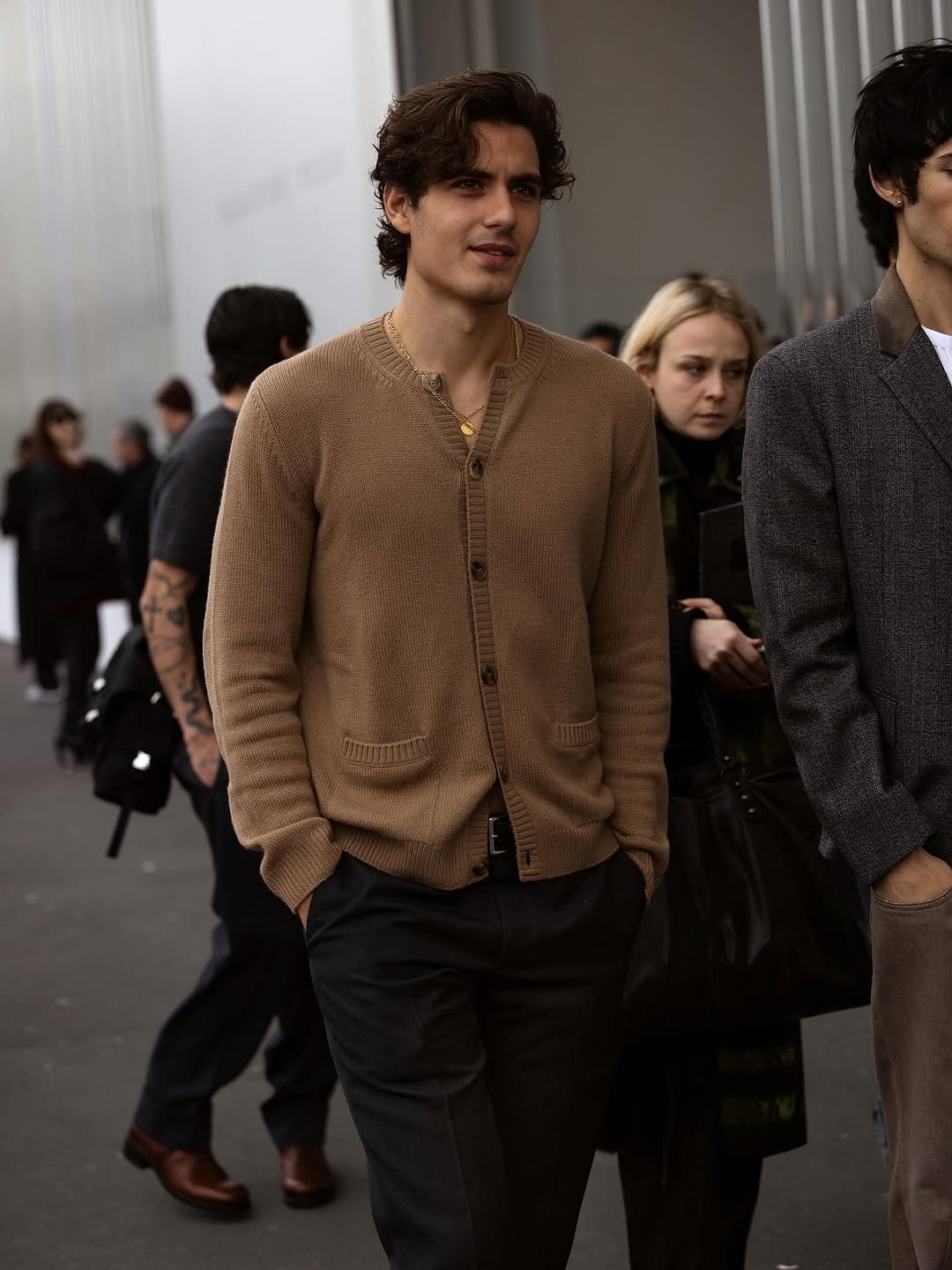 A tan knit cardigan is worn buttoned over dark tailored trousers with a black belt and layered gold necklaces.