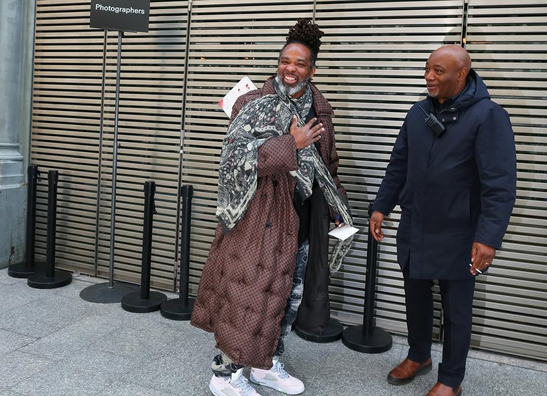 A layered street-style look with a long brown patterned coat, large printed scarf, dark layers, loose printed trousers, and pale sneakers.
