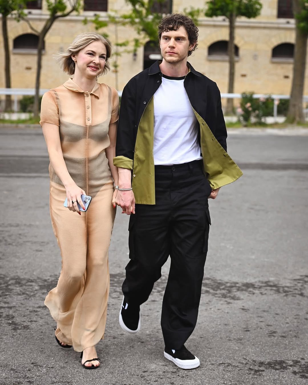 A couple walks in coordinated soft beige and black street-style looks, pairing a sheer polo jumpsuit with layered utility separates.