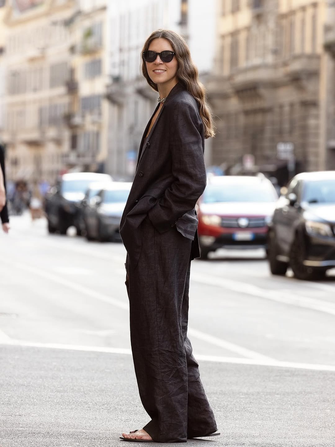 A dark relaxed linen suit is styled with black sunglasses, a short necklace, and flat sandals.