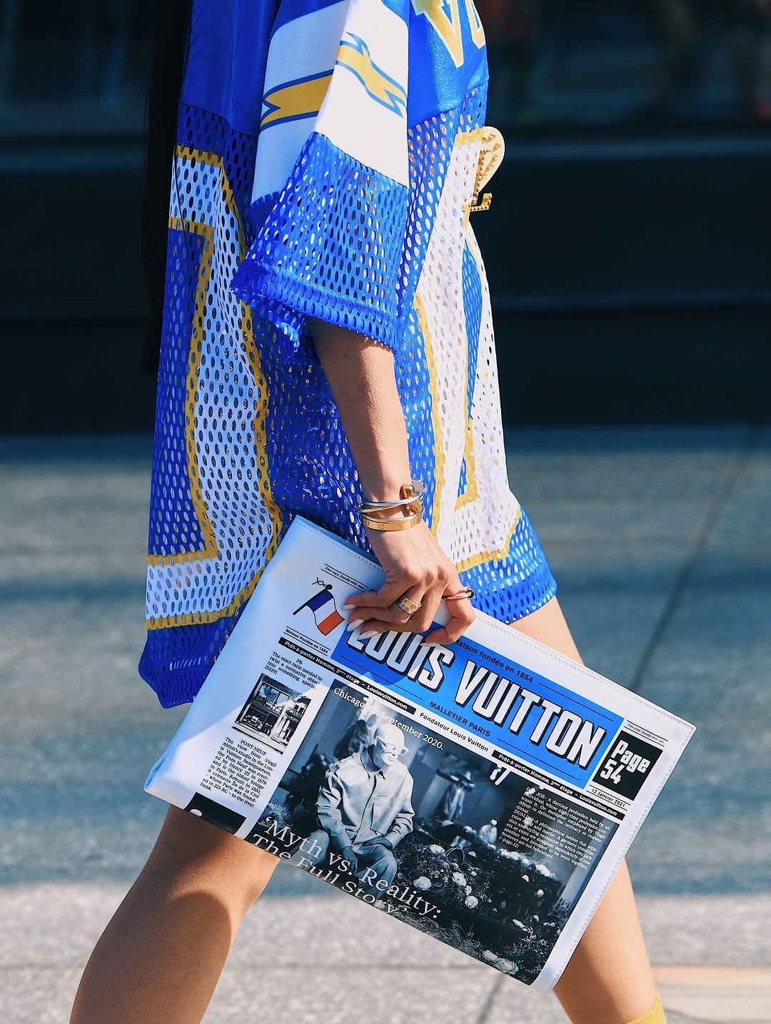 An oversized blue, white, and yellow mesh sports-jersey dress is styled with gold jewelry and a graphic Louis Vuitton newspaper clutch.