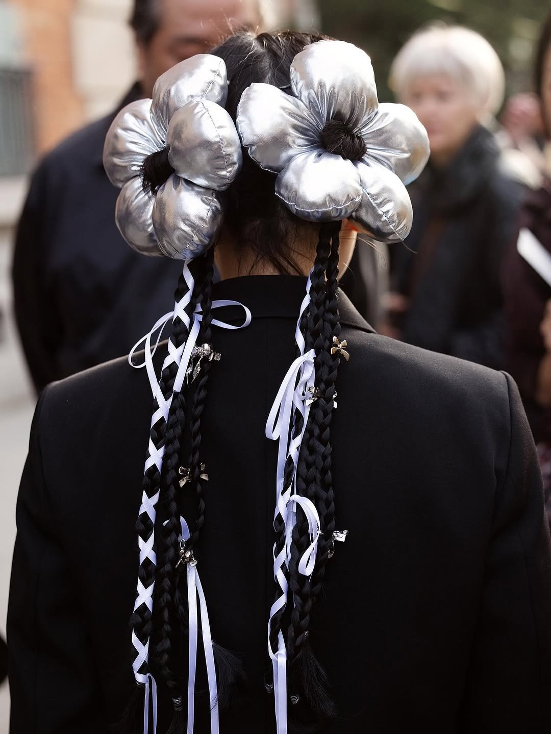 A black tailored jacket is styled with sculptural silver flower hair pieces, long braids, white ribbons, and small metallic charms.