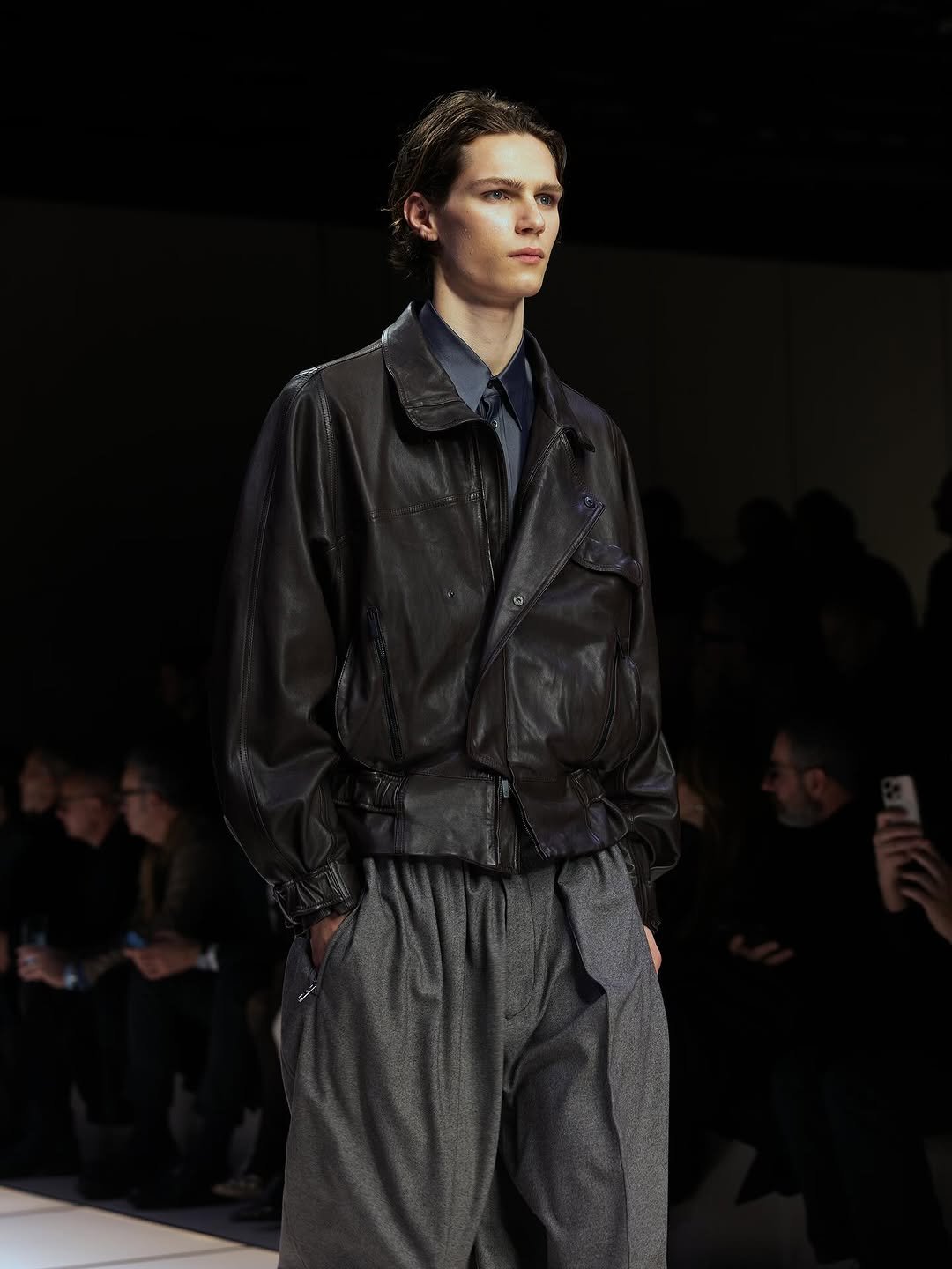 A black leather blouson jacket is layered over a dark shirt and tie with voluminous grey pleated trousers.