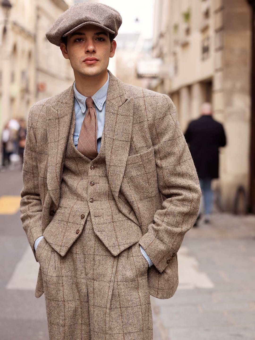 A tweed three-piece suit is styled with a light blue shirt, brown tie, and oversized flat cap.