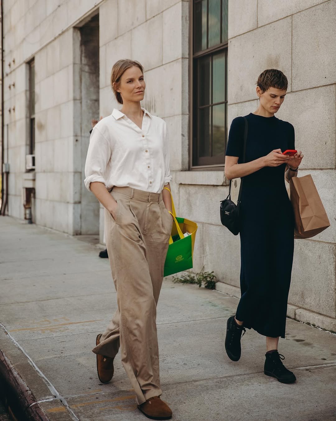 A white button-up shirt is tucked into beige pleated trousers with brown suede shoes and a bright green shopping bag.