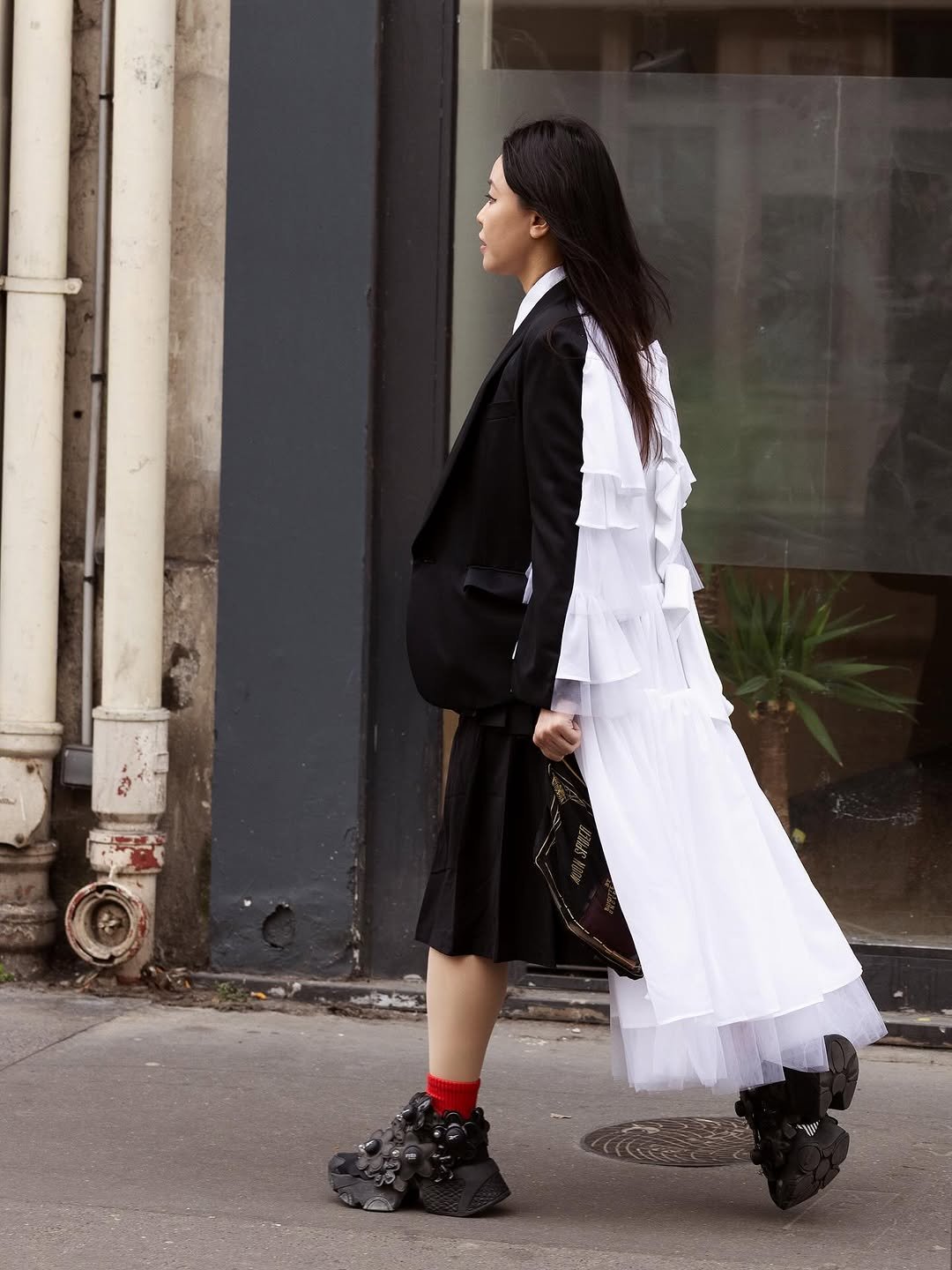 A black blazer-and-skirt look is disrupted by a long white ruffled back panel, red socks, sculptural black sneakers, and a black printed clutch.