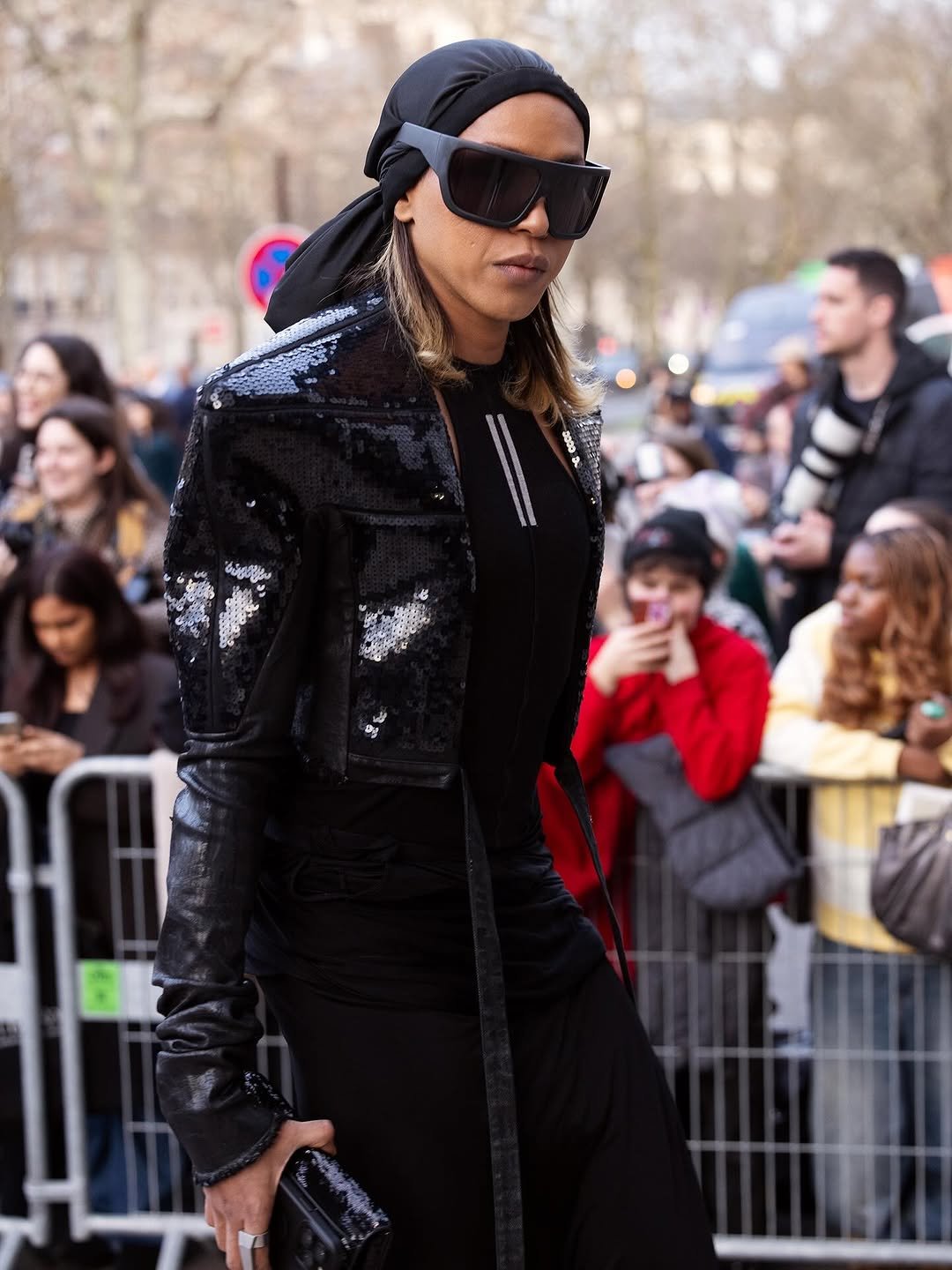 A black sequined cropped jacket is styled over a fitted black look with oversized angular sunglasses, a tied headscarf, and a glossy black clutch.