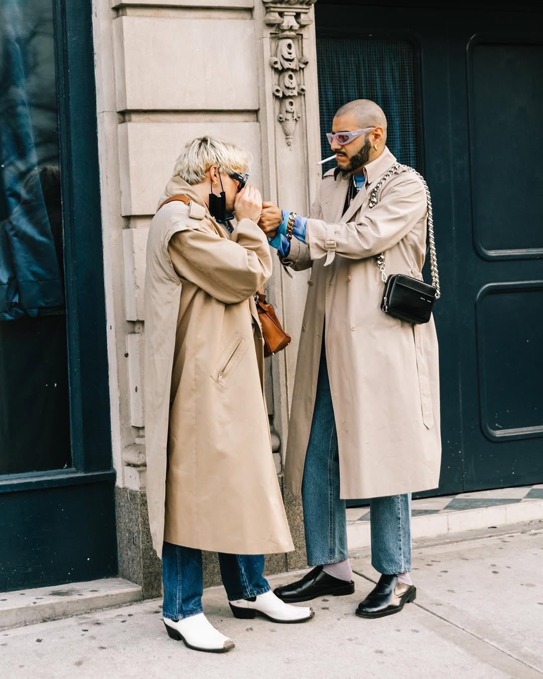Two wearers in beige trench coats style denim, ankle boots, sunglasses, and compact shoulder bags on a city sidewalk.