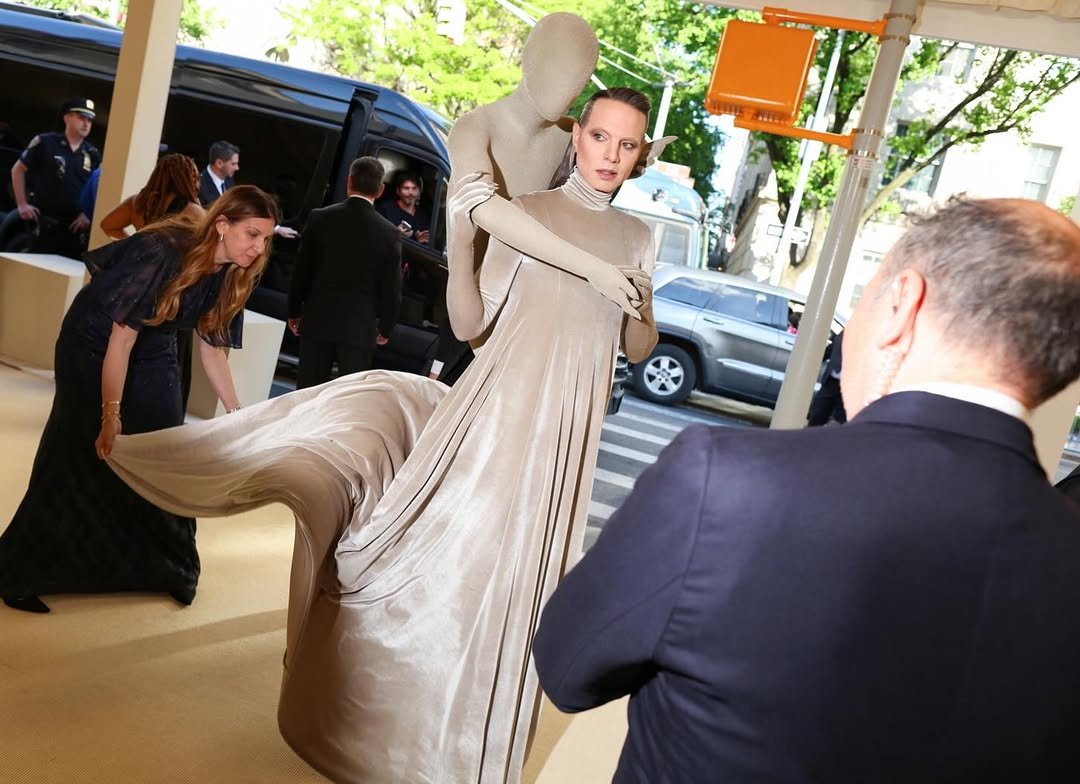 A champagne-beige sculptural draped gown is styled with matching gloves, a bodysuit-like hooded figure, and pointed ear detail.