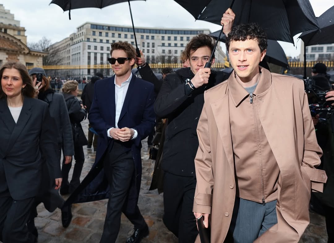 A taupe trench-style coat is layered over a tonal zip jacket, grey top, and blue-grey tailored trousers.