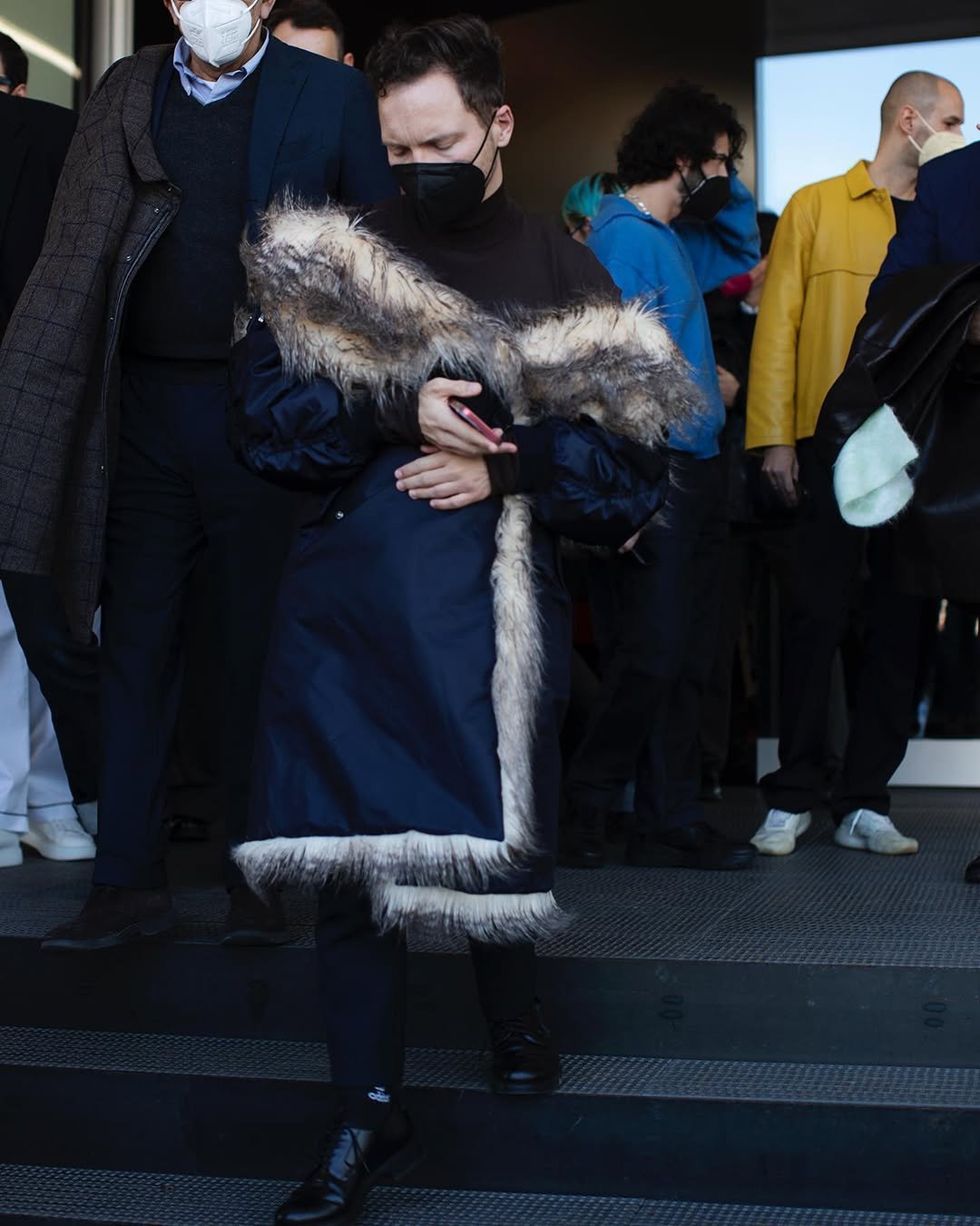 A dark turtleneck and trousers are styled with a dramatic navy coat edged in long shaggy faux fur and black lace-up shoes.