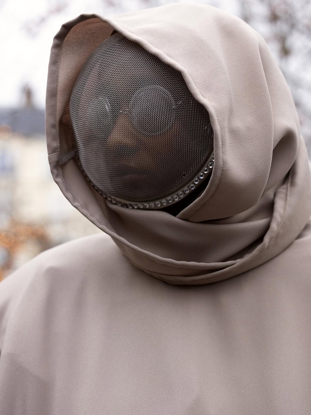 A taupe hooded draped garment frames a dark mesh face covering with a rhinestone-trimmed band.