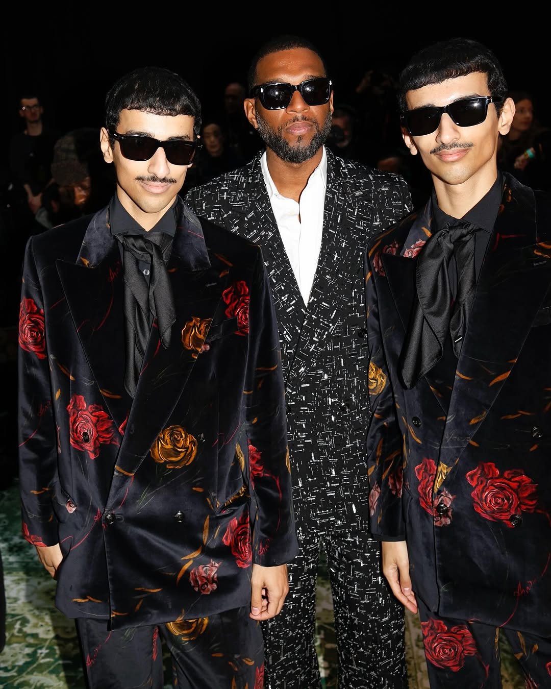 Three men wear dark statement suits with sunglasses, including matching black rose-print satin suits on the left and right and a black graphic-pattern suit in the center.
