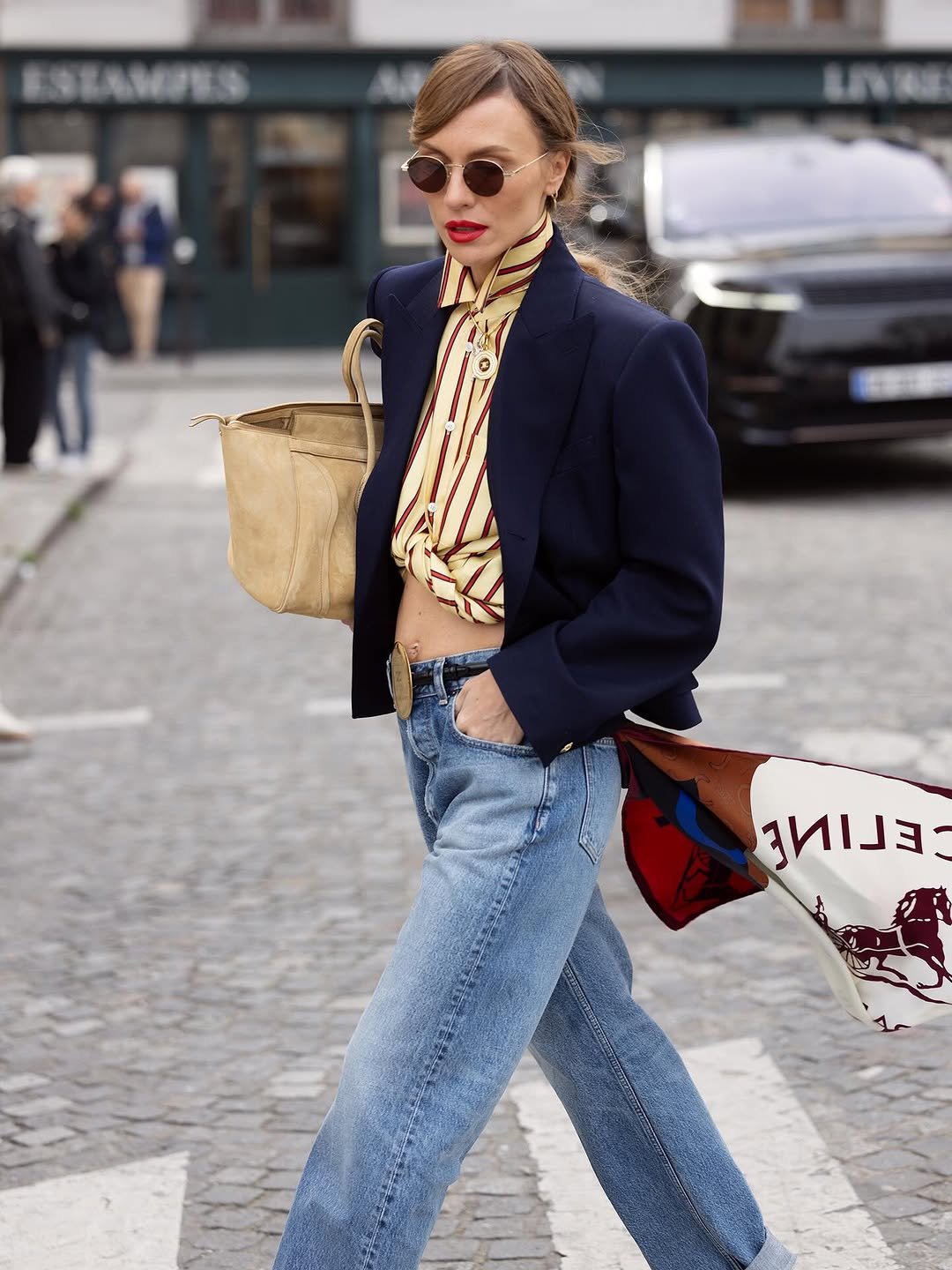 A navy blazer is styled over a knotted striped shirt with light-wash jeans, a tan belt, round sunglasses, and neutral handbags.