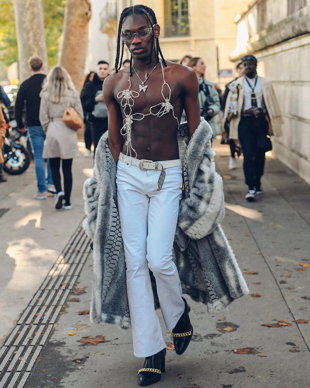 A shirtless street-style look pairs white flared trousers with a grey faux-fur coat, rhinestone body jewelry, a studded belt, and black chain-trim shoes.