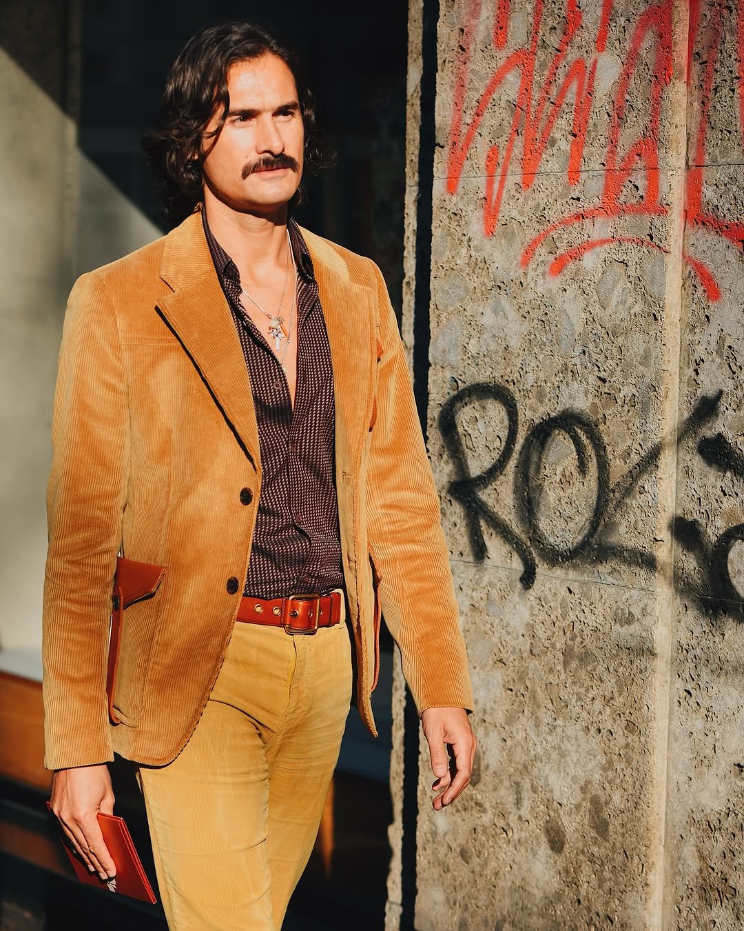 A mustard corduroy blazer and matching trousers are styled with a dark patterned open-collar shirt, red-brown belt, necklaces, and a red clutch.