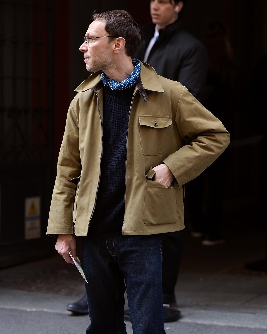 A khaki work jacket is worn over a dark knit sweater, dark jeans, and a blue dotted neck scarf.
