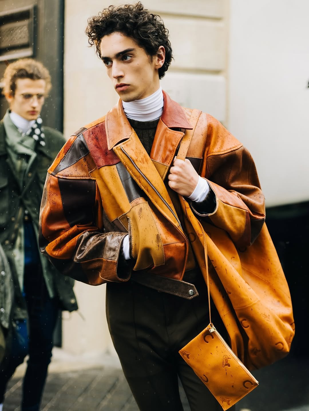 A patchwork tan leather jacket is layered over a white turtleneck and dark knit with dark trousers and a matching leather shoulder bag.