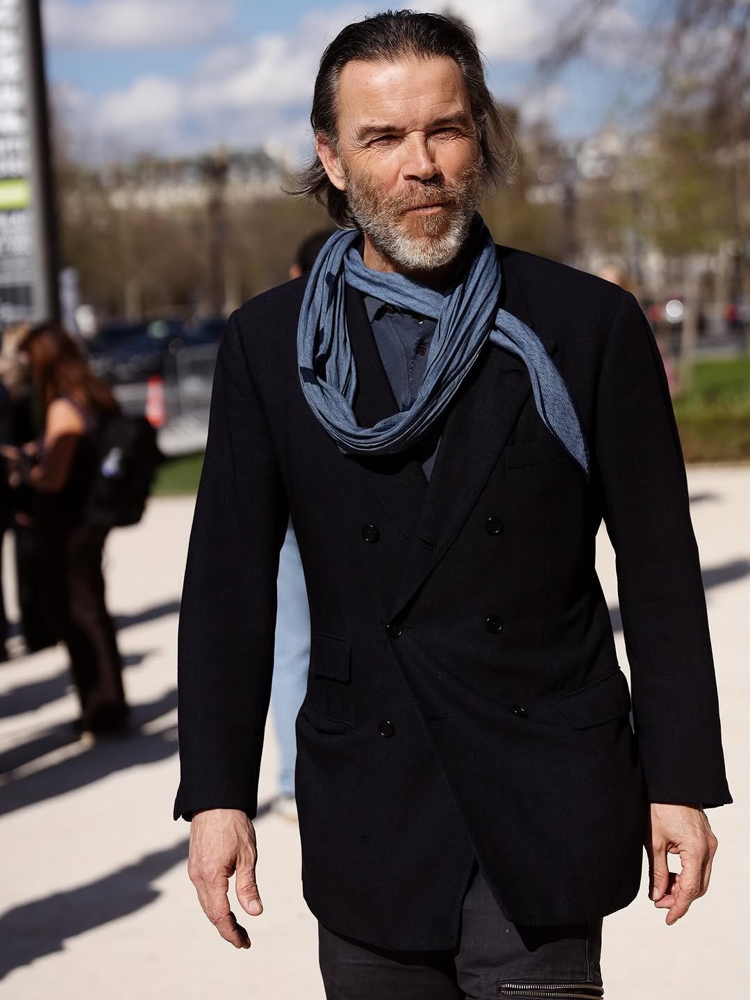 A black double-breasted blazer is worn over a dark shirt with a loosely wrapped blue scarf.