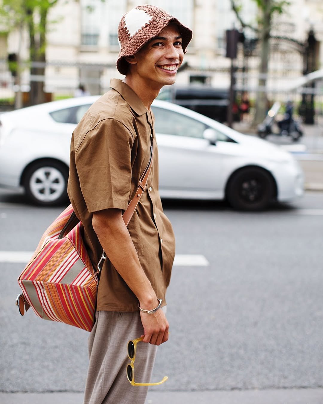 A brown short-sleeve shirt and taupe trousers are styled with a crochet bucket hat, striped shoulder bag, bracelet, and yellow sunglasses.