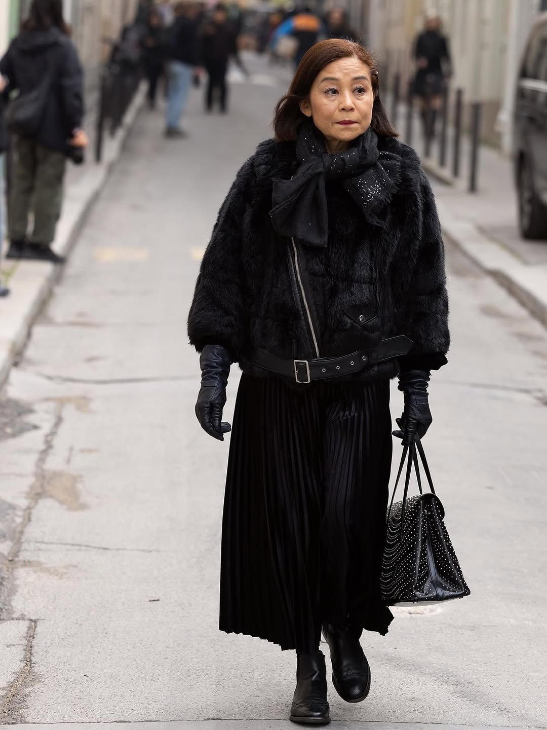 An all-black street-style look with a plush cropped jacket, sparkling scarf, pleated skirt, leather gloves, studded bag, and black boots.