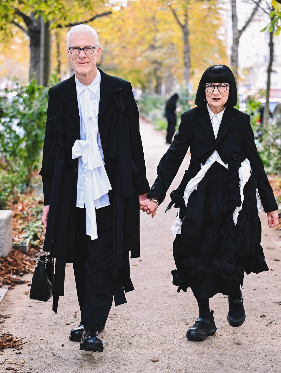 Two wearers in black-and-white layered tailoring walk outdoors with long coats, crisp shirts, sculptural fabric ties, and chunky black shoes.
