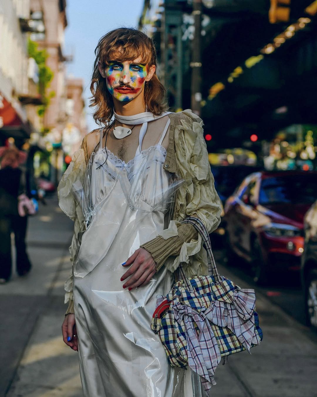 An avant-garde layered white slip look is styled with a sheer crumpled jacket, plastic overlay, bold face paint, and a patchwork plaid ruffle bag.