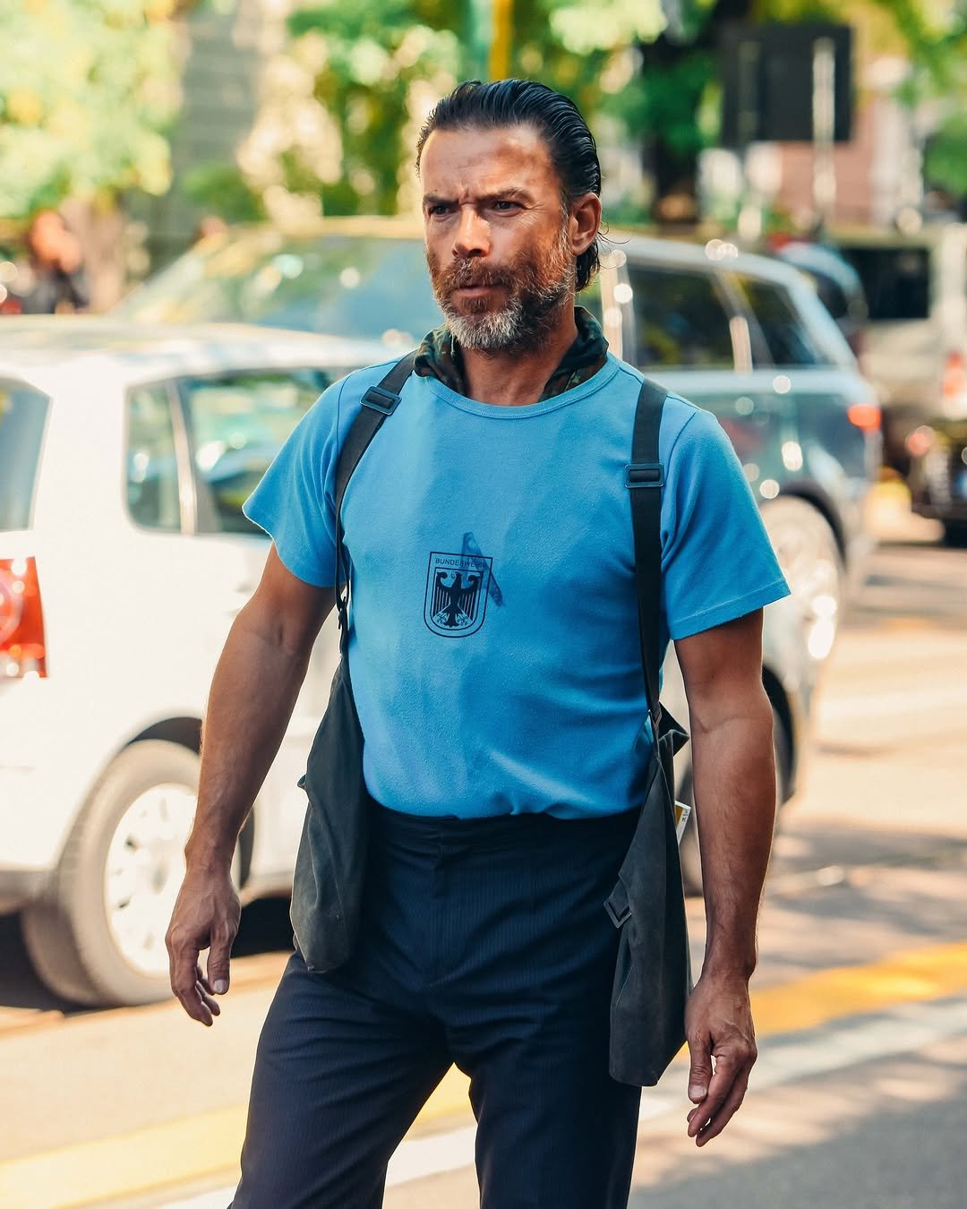 A blue graphic T-shirt is tucked into dark high-waisted trousers with black shoulder straps and a dark outer layer hanging behind.