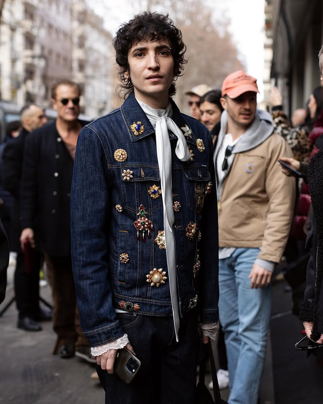 A dark embellished denim jacket is styled over a white neck-tie blouse with lace cuffs and dark trousers.