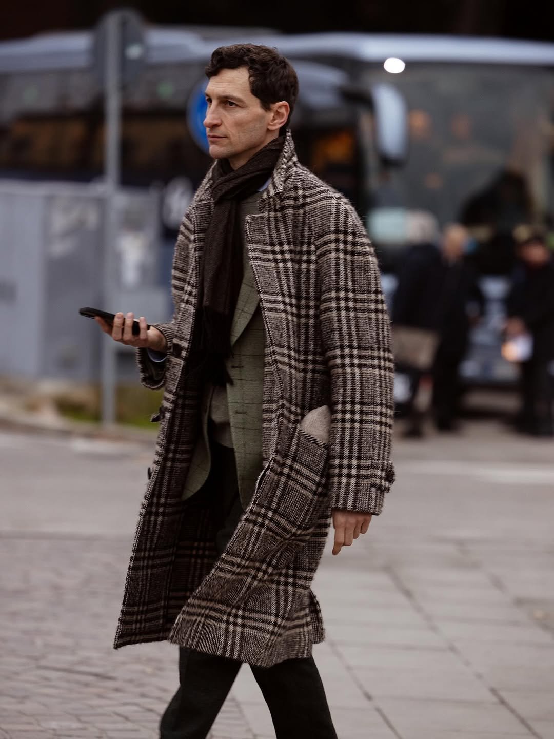A checked overcoat is layered over a muted green blazer, dark scarf, and black trousers.