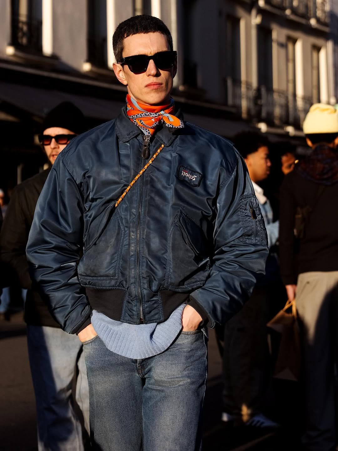 A navy bomber jacket is layered over a pale blue ribbed knit with blue jeans, black sunglasses, and an orange patterned neck scarf.