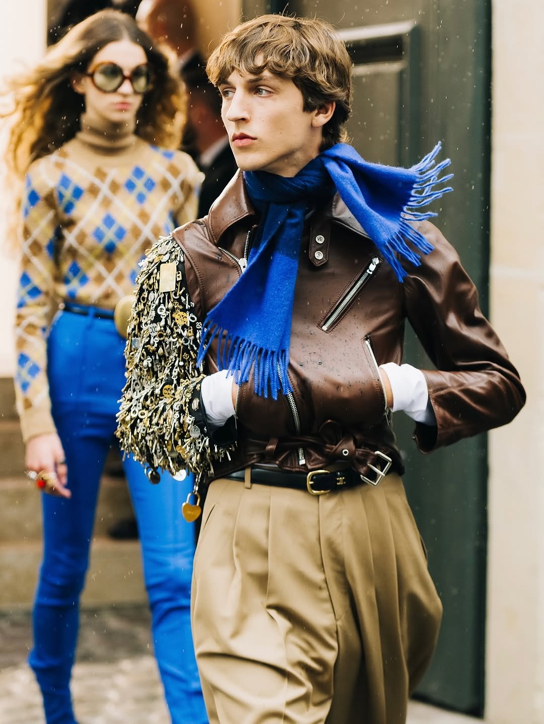 A brown leather moto jacket is styled with a cobalt fringed scarf, beige pleated trousers, white gloves, and an embellished shoulder bag.