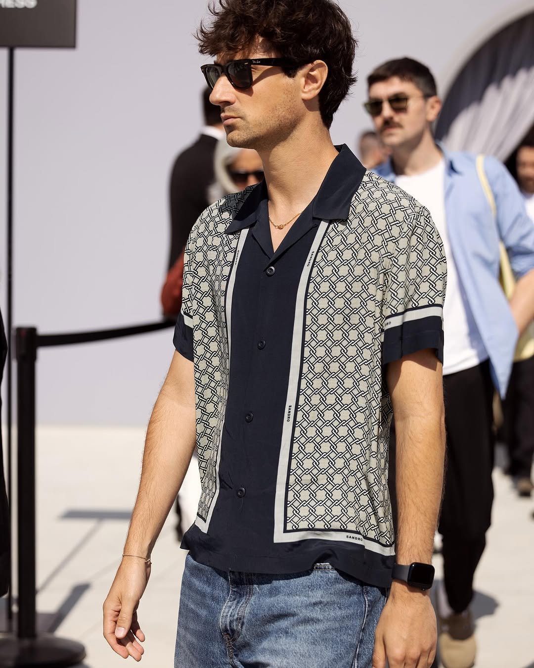 A patterned short-sleeve camp-collar shirt is styled with light blue denim, black sunglasses, a fine gold necklace, bracelet, and smartwatch.