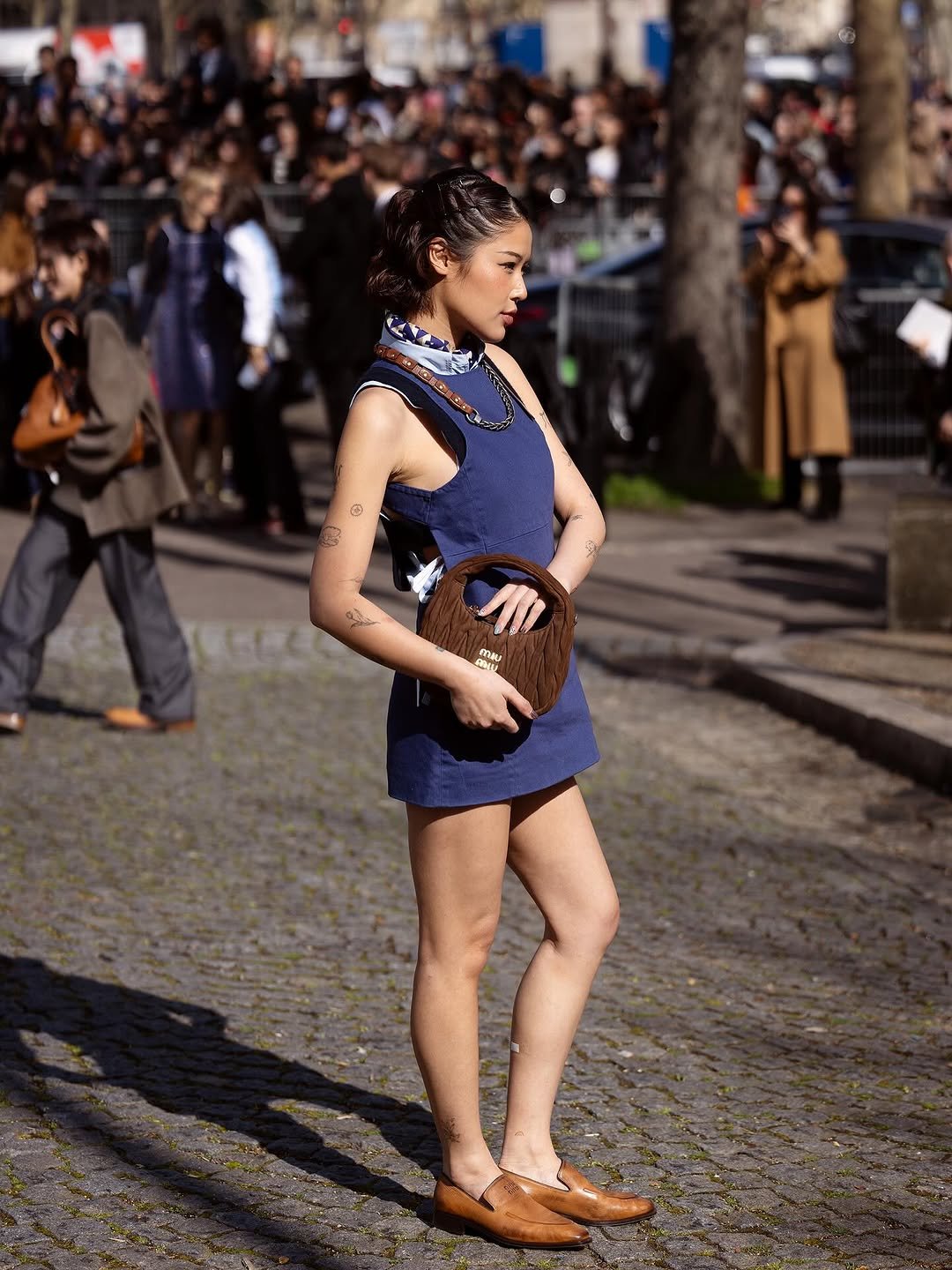 A fitted navy sleeveless mini dress is styled with a printed neck scarf, brown loafers, and a ruched brown top-handle bag.