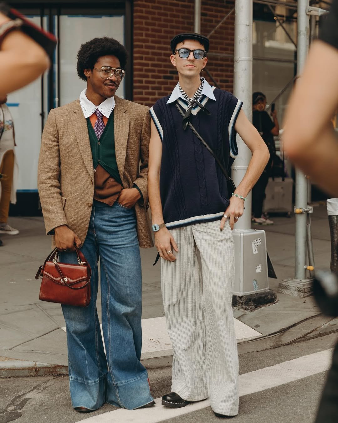 Two men wear layered preppy street-style looks with tailoring, knitwear, wide trousers, denim, and visible accessories.