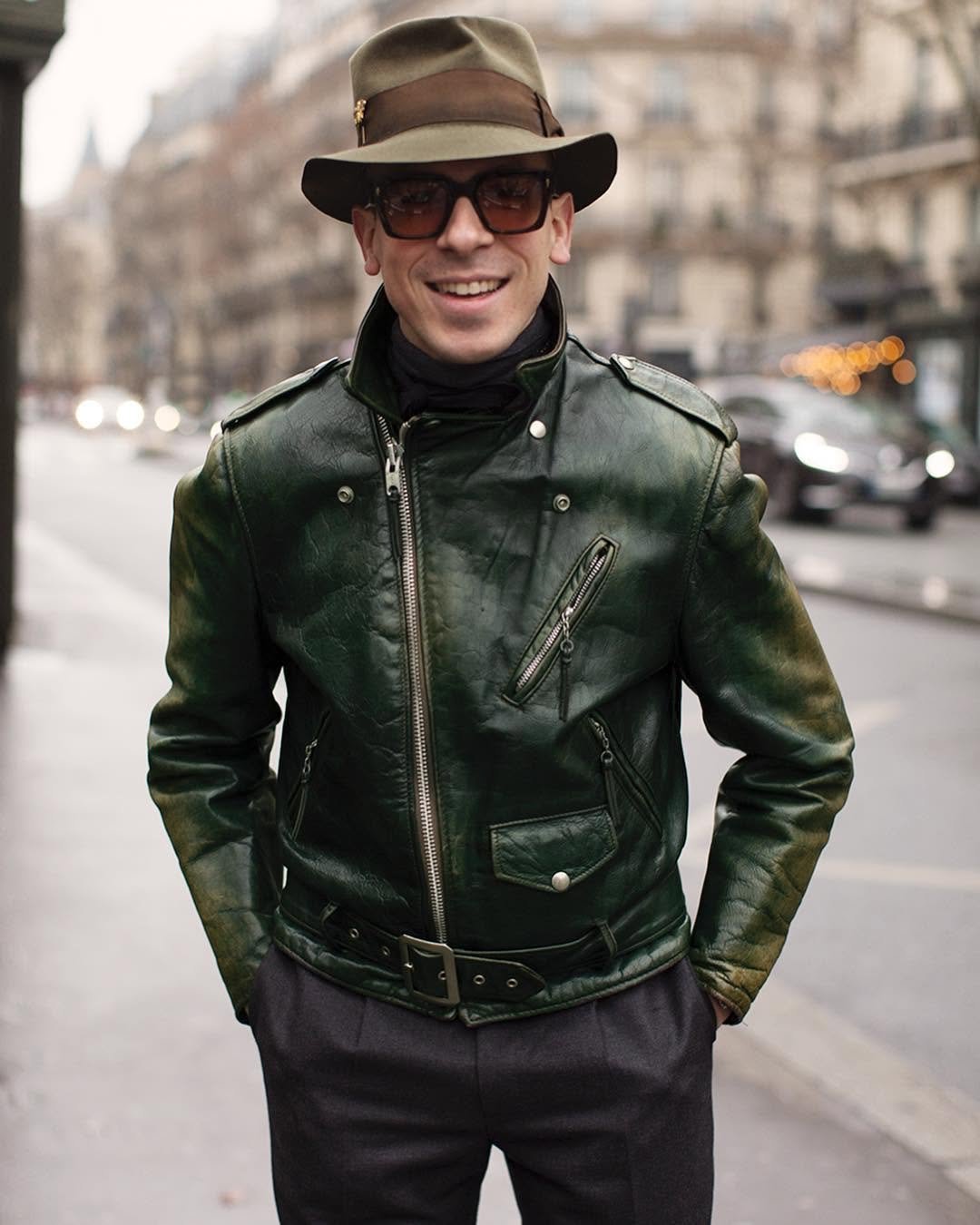 A dark green leather biker jacket is styled with tailored dark trousers, a wide-brim hat, tinted sunglasses, and a black neck layer.