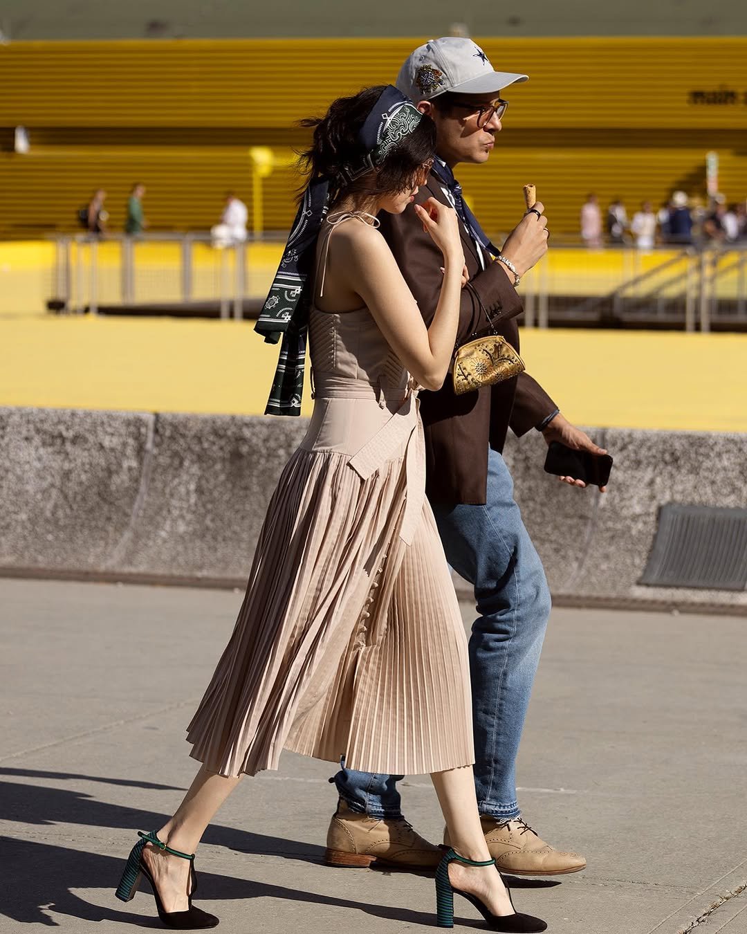 A beige strappy pleated midi dress is styled with a dark patterned headscarf, sunglasses, and black ankle-strap heels with green accents.
