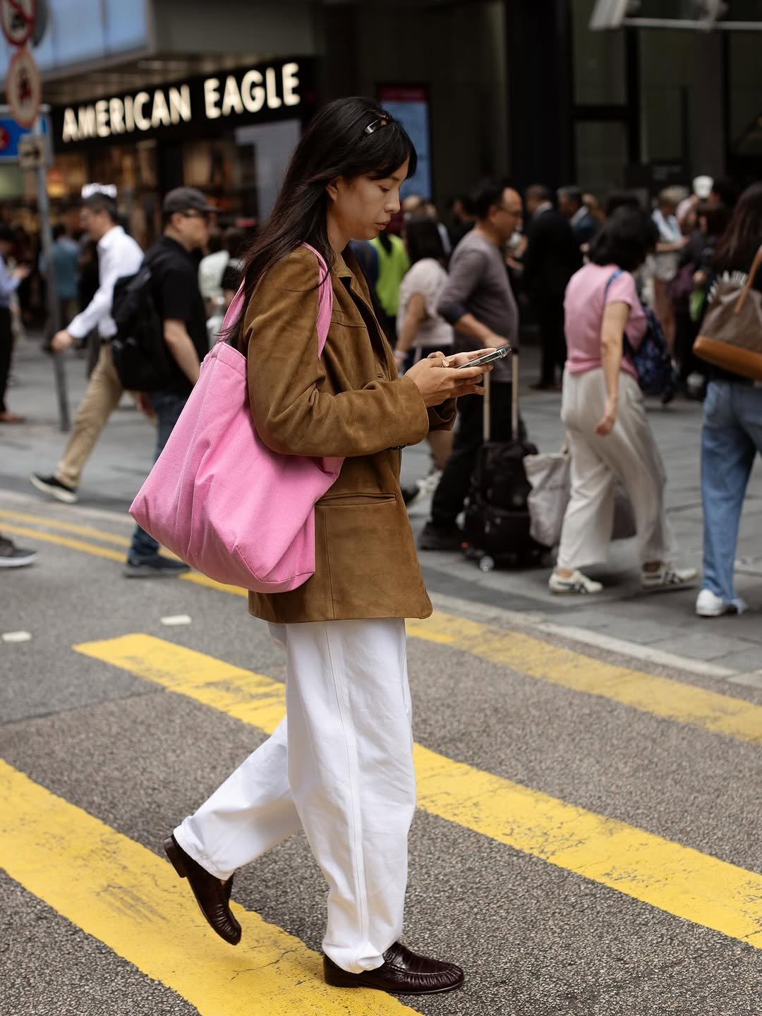 A brown suede-like jacket is styled with loose white trousers, dark loafers, and a large pink shoulder tote.
